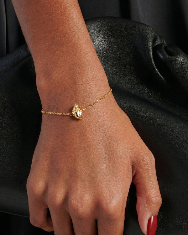 Gold bracelet featuring an anatomical heart pendant styled on a model holding a black leather clutch