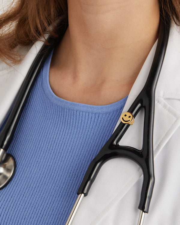 Gold plated smiley face stethoscope charm clipped to black stethoscope tubing worn over blue top and white coat