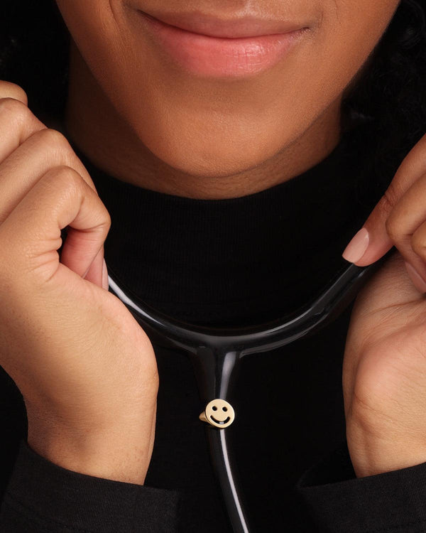 Gold plated smiley face stethoscope charm attached to black stethoscope tubing, held near the neck